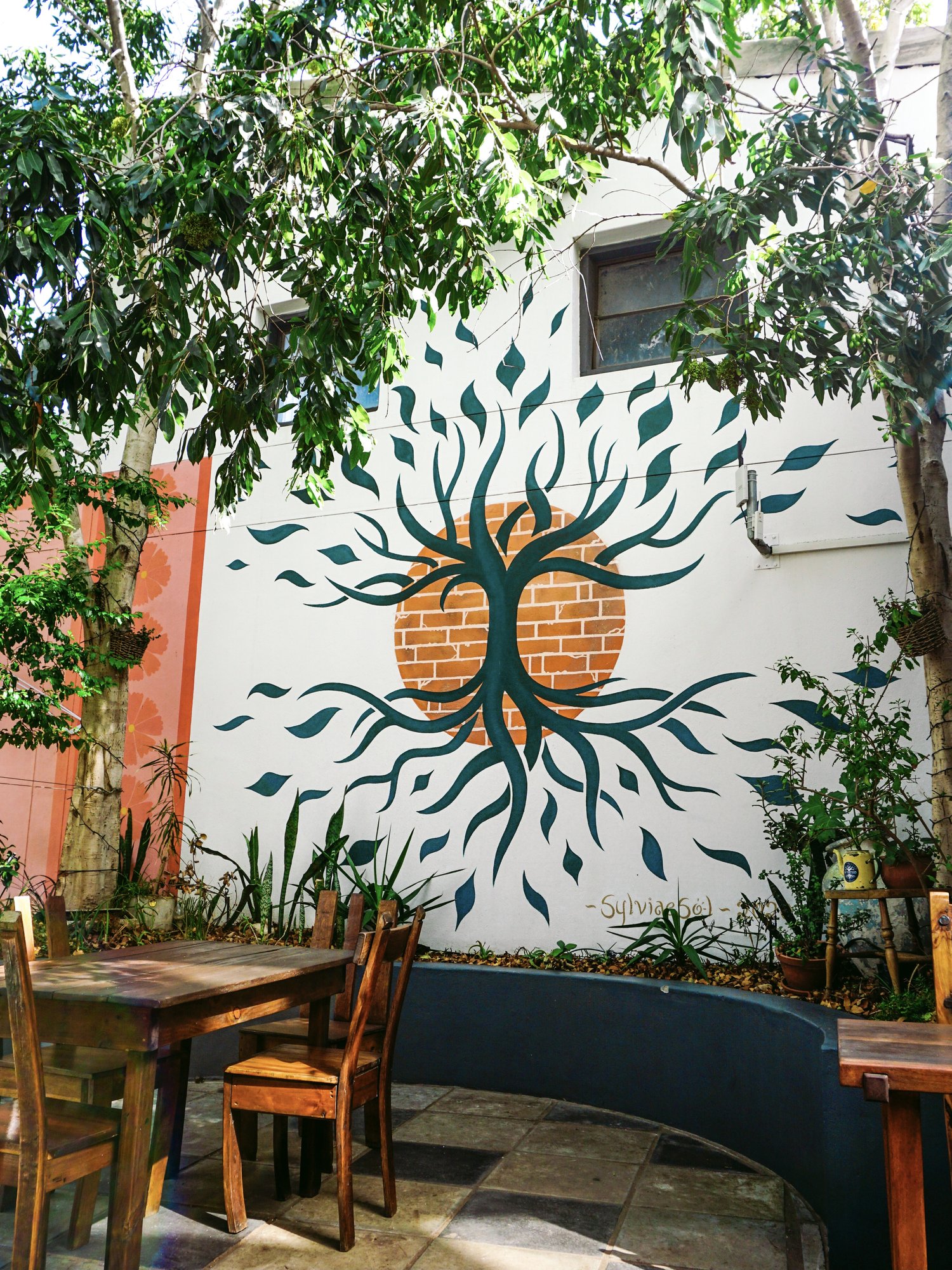 The Courtyard Cafe tree-of-life mural framed by garden foliage and cafe seating.